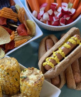 a bbq spread with corn, hot dogs, chips, and veggies