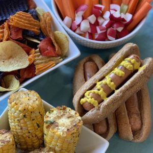 a bbq spread with corn, hot dogs, chips, and veggies