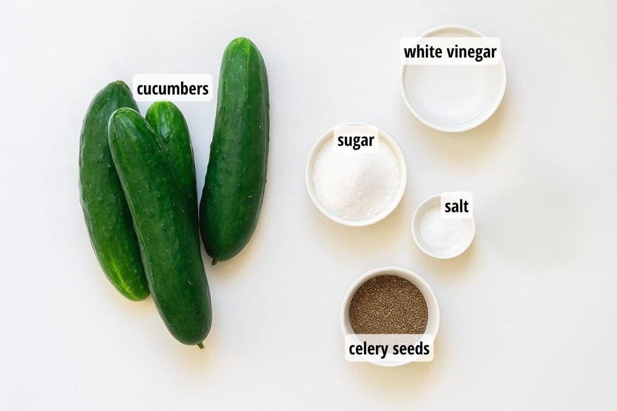 A white counter top with four cucumbers and four small bowls of labeled ingredients needed to make sweet pickles.