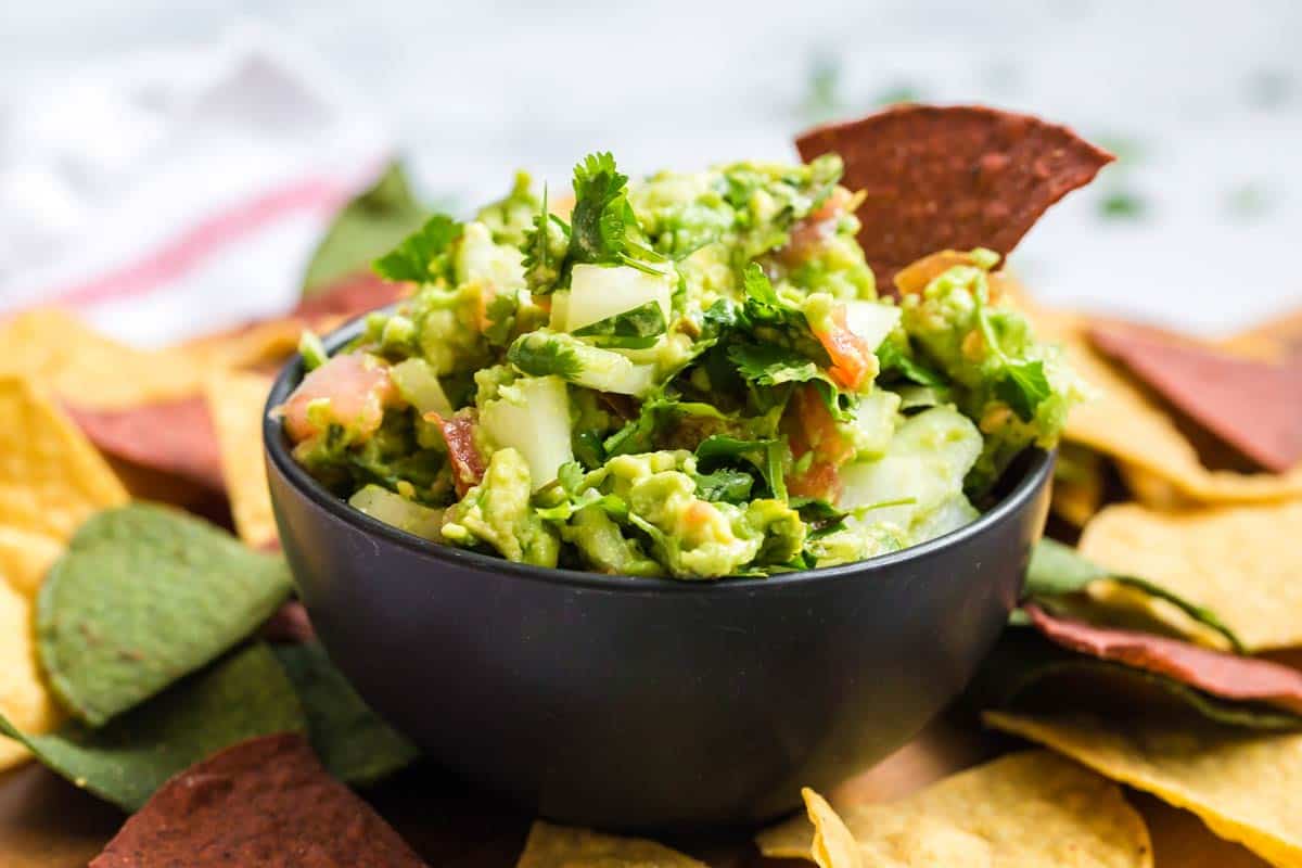 Black bowl with homemade guacamole. There are red, green, and yellow tortilla chips around the bowl. 