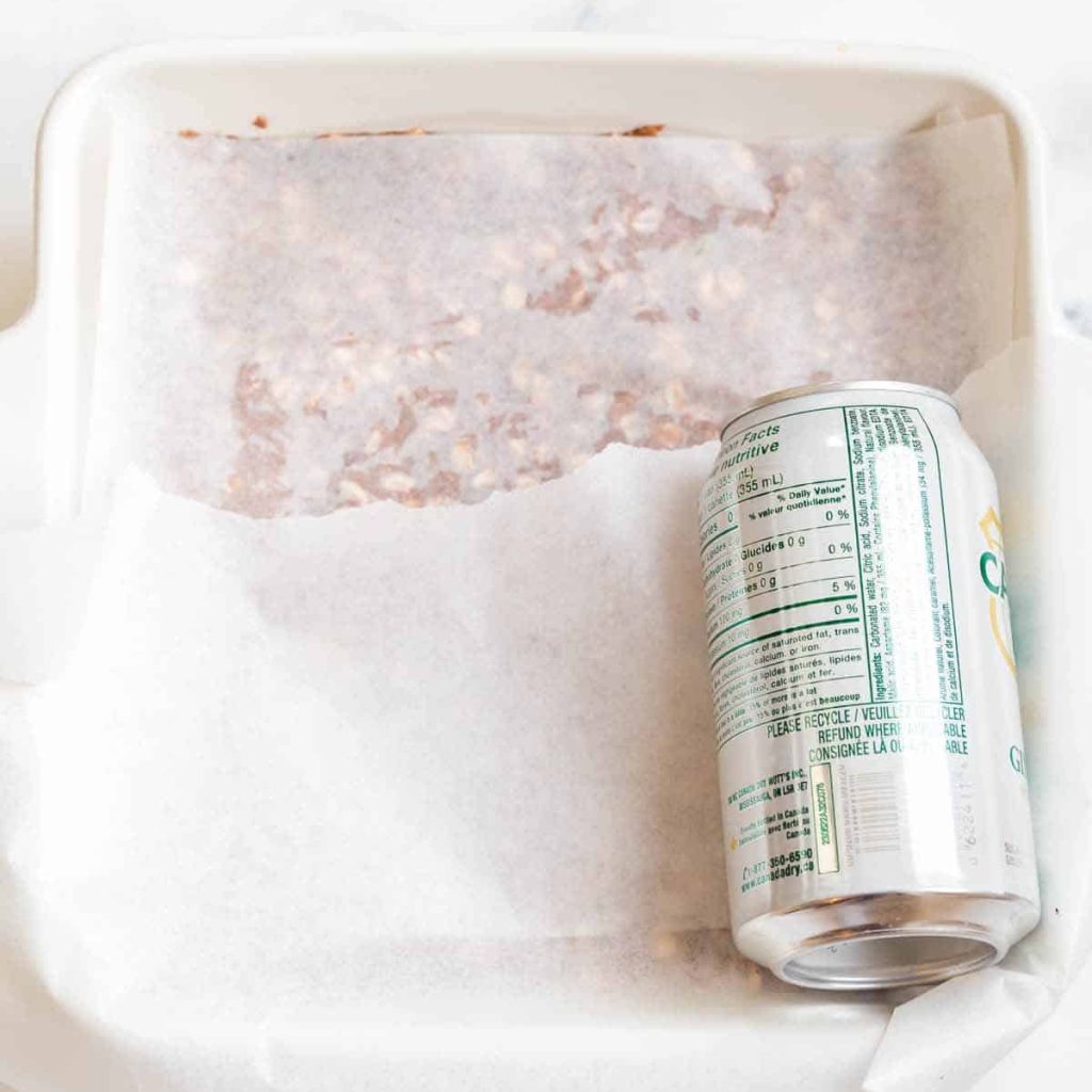 peanut butter granola bar mixture in a pan, under a piece of parchment paper, being smashed by a can of soda