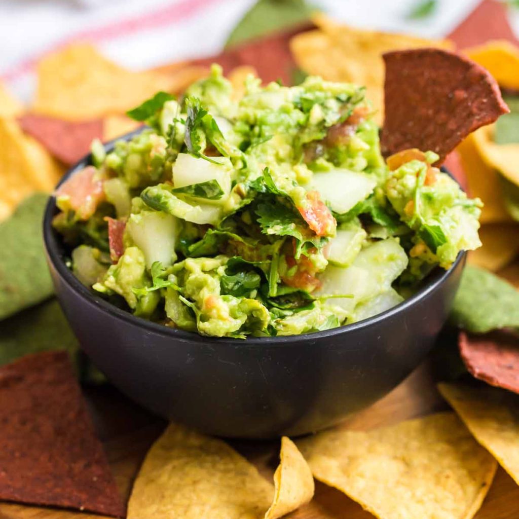 fresh homemade guacamole in a black bowl. THere is a dark red tortilla chip sticking out of the guacamole, and there are more tortilla chips laid out around the bowl.