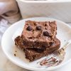 horizontal image of two black bean brownies stacked on a white plate with a fork next to them. Countertop is white, as is a pan of brownies in the background.