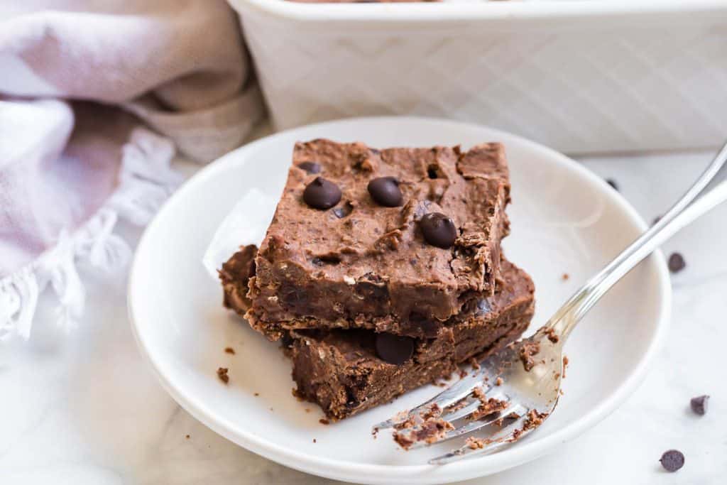 horizontal image of two black bean brownies stacked on a white plate with a fork next to them. Countertop is white, as is a pan of brownies in the background.