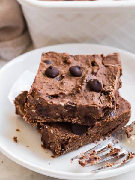 horizontal image of two black bean brownies stacked on a white plate with a fork next to them. Countertop is white, as is a pan of brownies in the background.