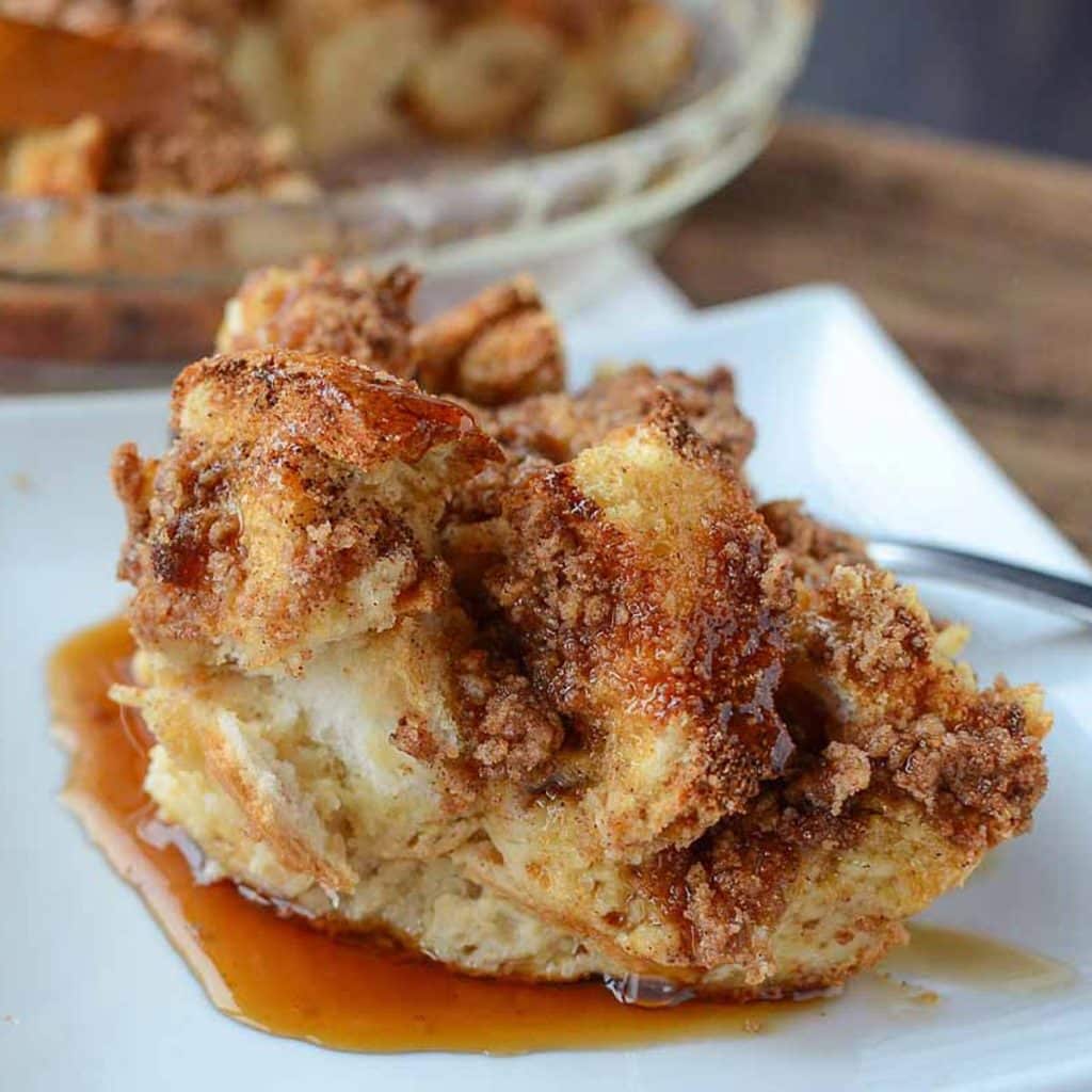 A white square plate with a slice of dinner roll french toast casserole topped with a cinnamon sugar strudel and maple syrup.