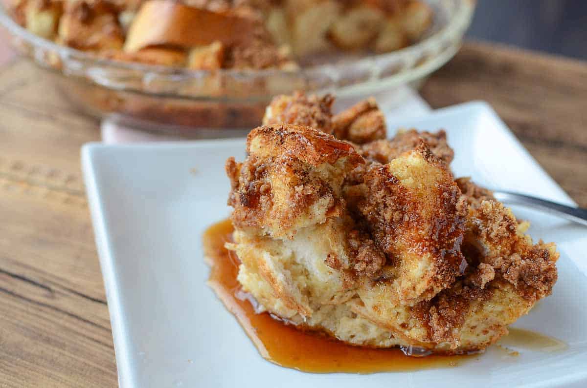 A white square plate with a slice of dinner roll french toast casserole topped with a cinnamon sugar strudel and maple syrup.