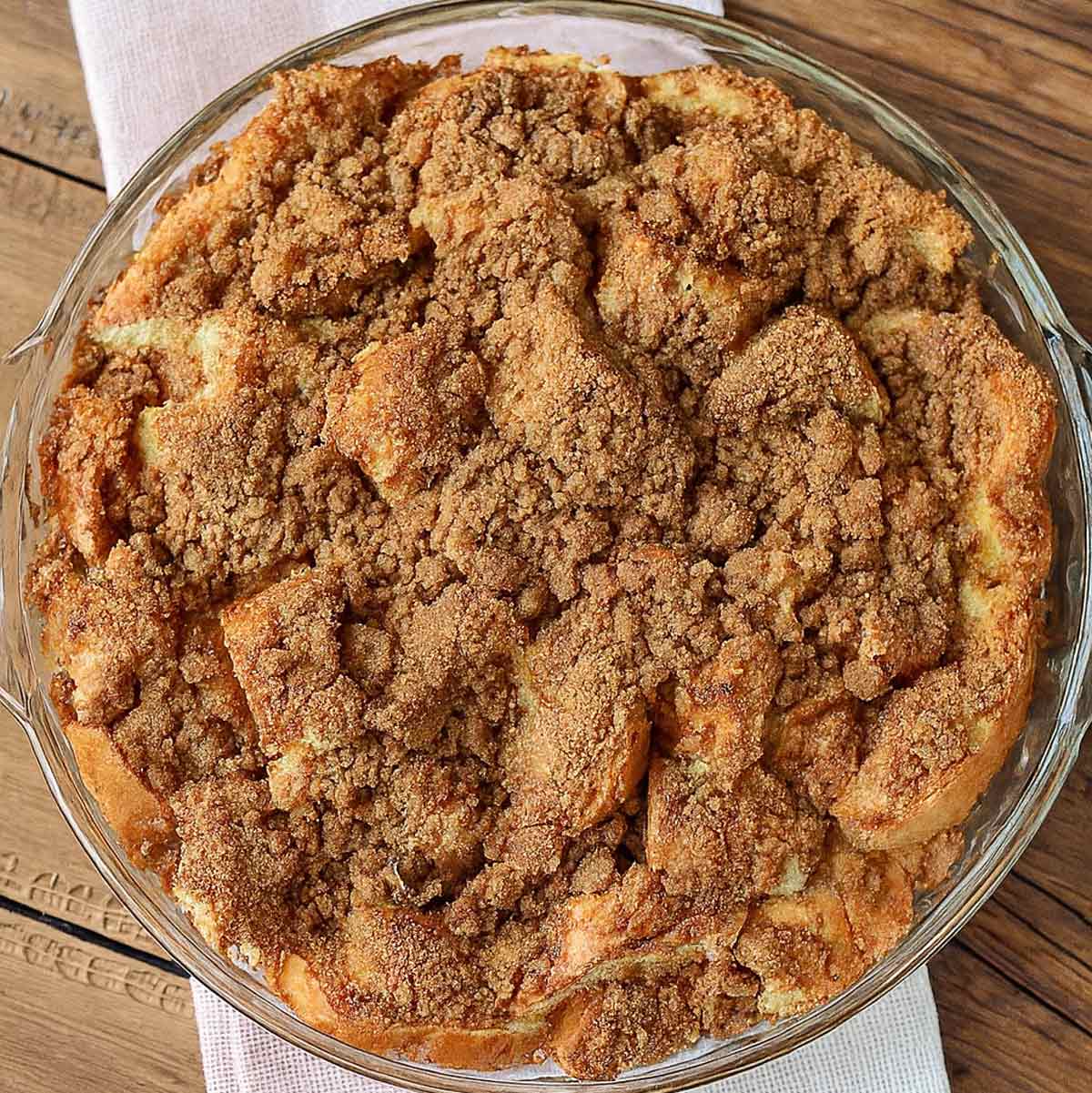 A circular baking dish filled with french toast casserole and topped with a cinnamon strudel, sitting on a folded red and white napkin on a wooden table.