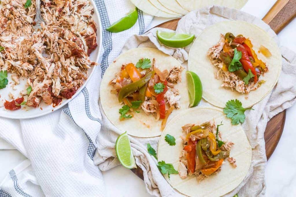 Three chicken fajitas are arranged on a wooden platter. Garnished with lime wedges and cilantro. In the upper left hand corner of the picture, there is a plate with remaining chicken fajitas. 
