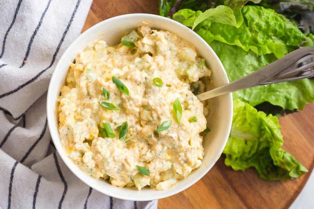 White bowl filled with healthy egg salad that is a slight yellow color. The egg salad is garnished with sliced green onions. Below the bowl is a white and grey striped napkin, and a wooden cutting board. Fresh chopped lettuce is to the right of the bowl.