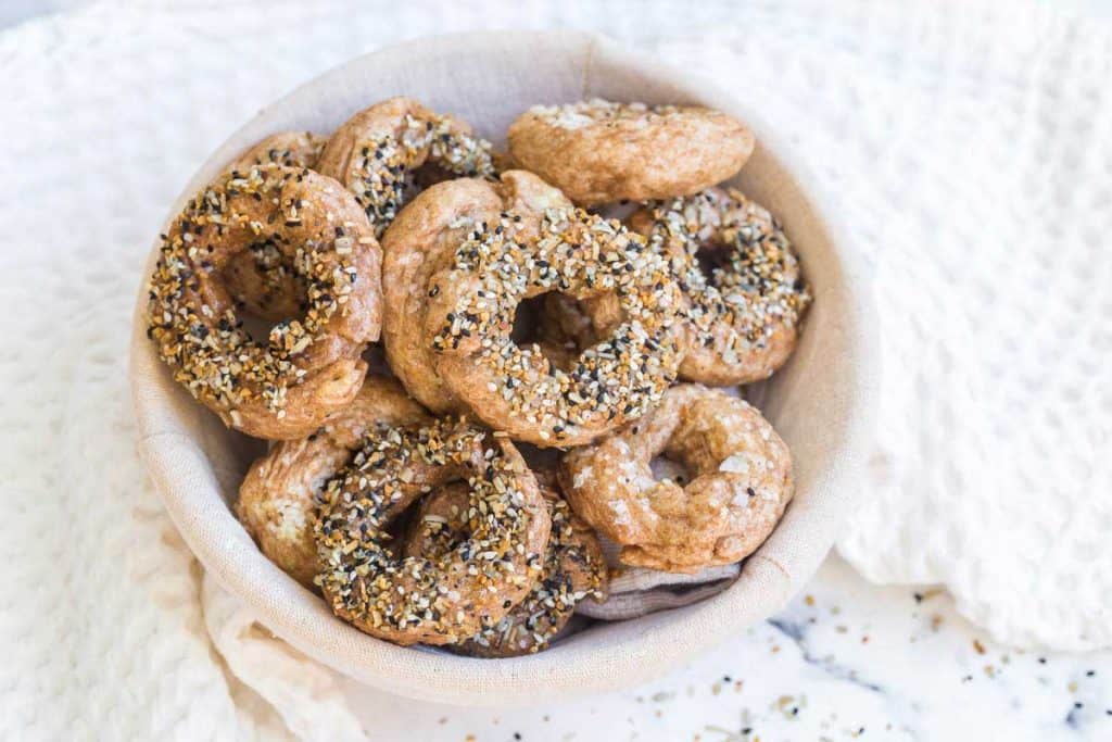Image of a bowl of whole wheat bagels sitting on top of a piece of white linen cloth. The bagels are topped with Everything But the Bagel Seasoning. 