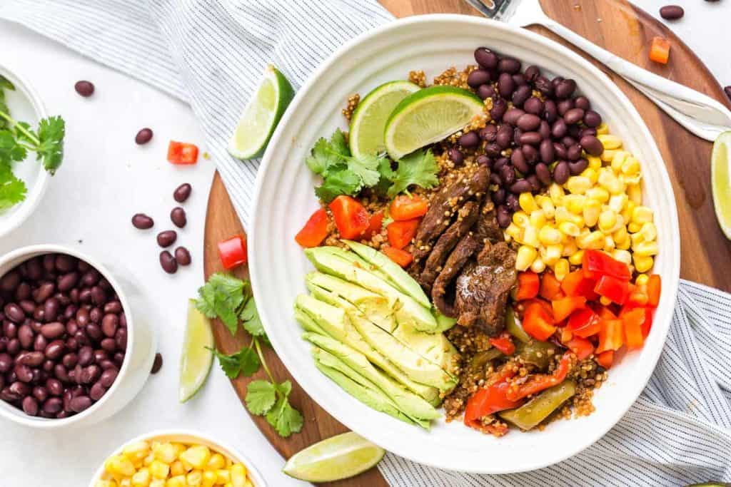 Image of steak bowl garnished with avocado, corn, diced tomato, black beans, and lime wedges.