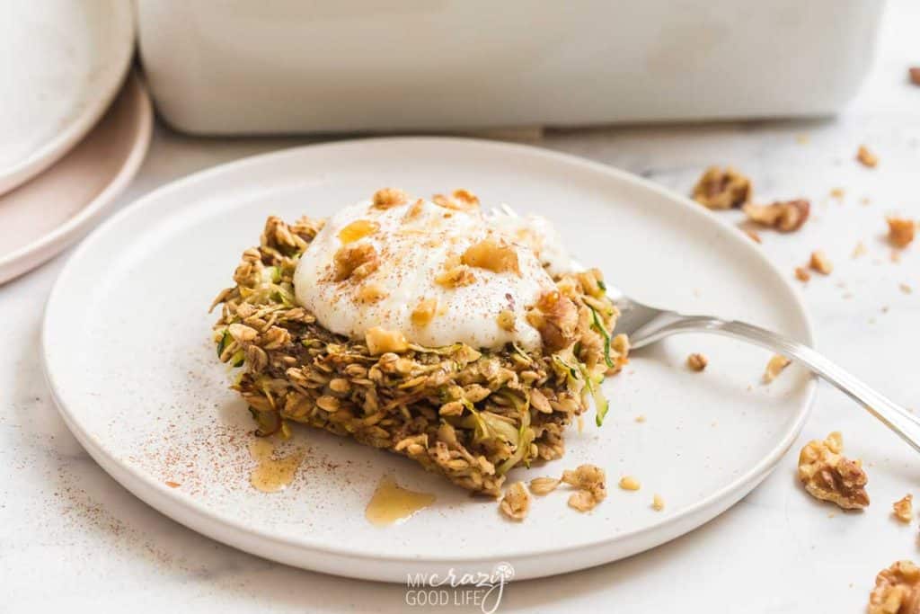 Image of pice of zucchini oatmeal bar on a white plate. The bar is garnished with greek yogurt, maple syrup, and walnuts.  