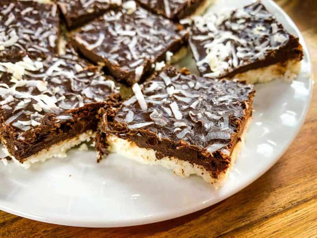 On top of a wooden table there is a white plate with several coconut bars sitting on top. The coconut bars are made with a coconut base, topped with a chocolate layer, and sprinkled with shredded coconut. 