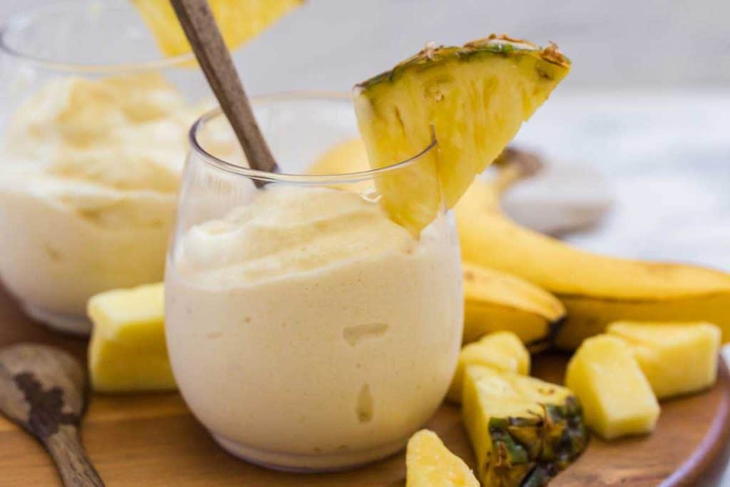 A tasty dole whip treat is in a clear small drinking glass. The glass is filled almost to the top. The glass is garnished with a wedge of pineapple sitting on the rim. IN the background are various pieces of pineapple, and a few bananas.