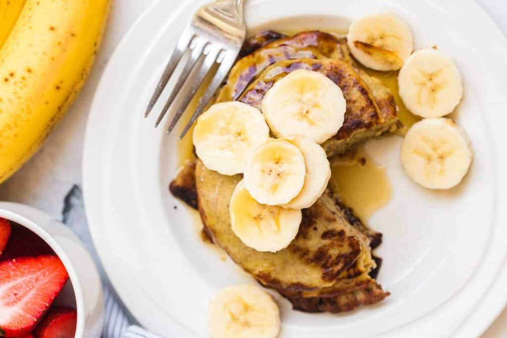 Image of 2 ingredient banana pancakes stacked on top of white plate. On top of the pancake stack are sliced banana pieces and a drizzle of maple syrup. A silver fork is resting in the upper left hand corner of the plate. Next to the plate is a banana and a small bowl of strawberries. 
