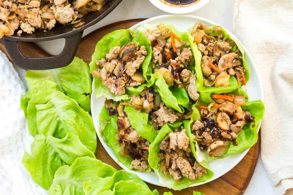 A black cast iron skillet with ground turkey and seasonings is located in the upper left hand of the photo. In the center, there is a white plate filled with six turkey lettuce wraps. 