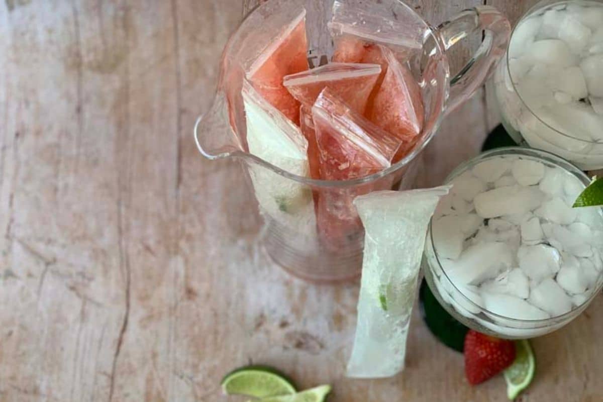 A wooden counter top with a clear pitcher full of margarita popsicles and some lime wedges.