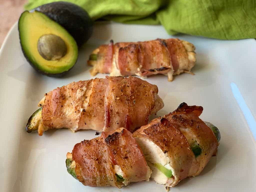 Image of three avocado stuffed chicken pieces sitting on white plate. The pieces are each wrapped in crispy cooked bacon. In the back of the plate, there is half of an avocado with the pit inside, and a green cloth napkin. 