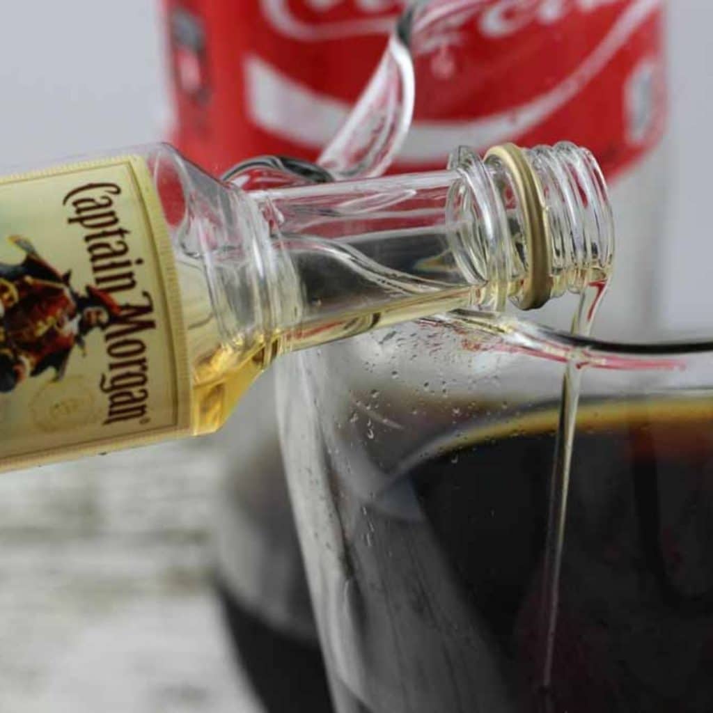 A small bottle of rum being poured into a glass with coca-cola in it.