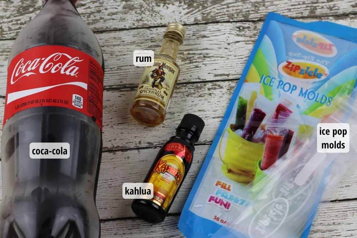 A rustic wooden table top with bottles of coca-cola, rum, and kahlua, and a package of popsicle molds.
