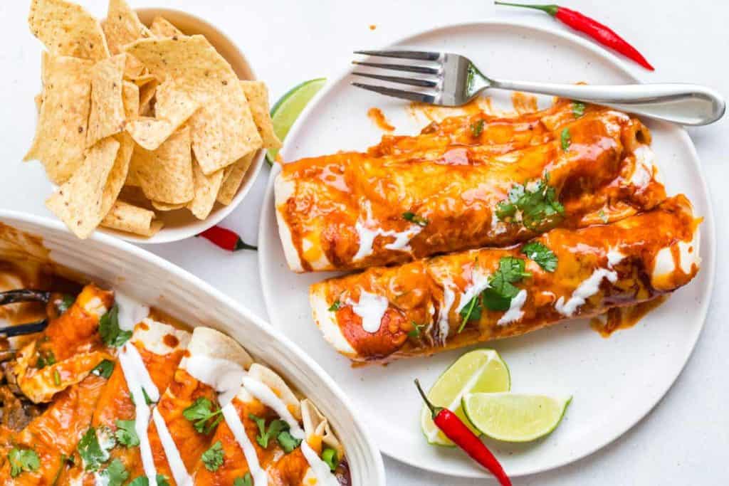 Image of two enchiladas on a white plate with a fork on plate. On the left, next to the plate is a small bowl of tortilla chips. Below the bowl is a serving dish of more enchiladas. The enchiladas are covered in a red sauce and garnished with a white drizzle, cilantro, and lime wedges. 
