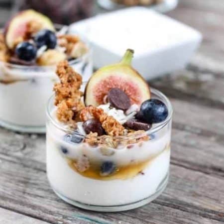A glass cup with a white yogurt mixture on the bottom and topped with granola, blueberries, a piece of fig, and chocolate.