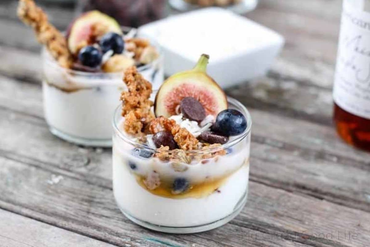 A glass cup with a white yogurt mixture on the bottom and topped with granola, blueberries, a piece of fig, and chocolate.