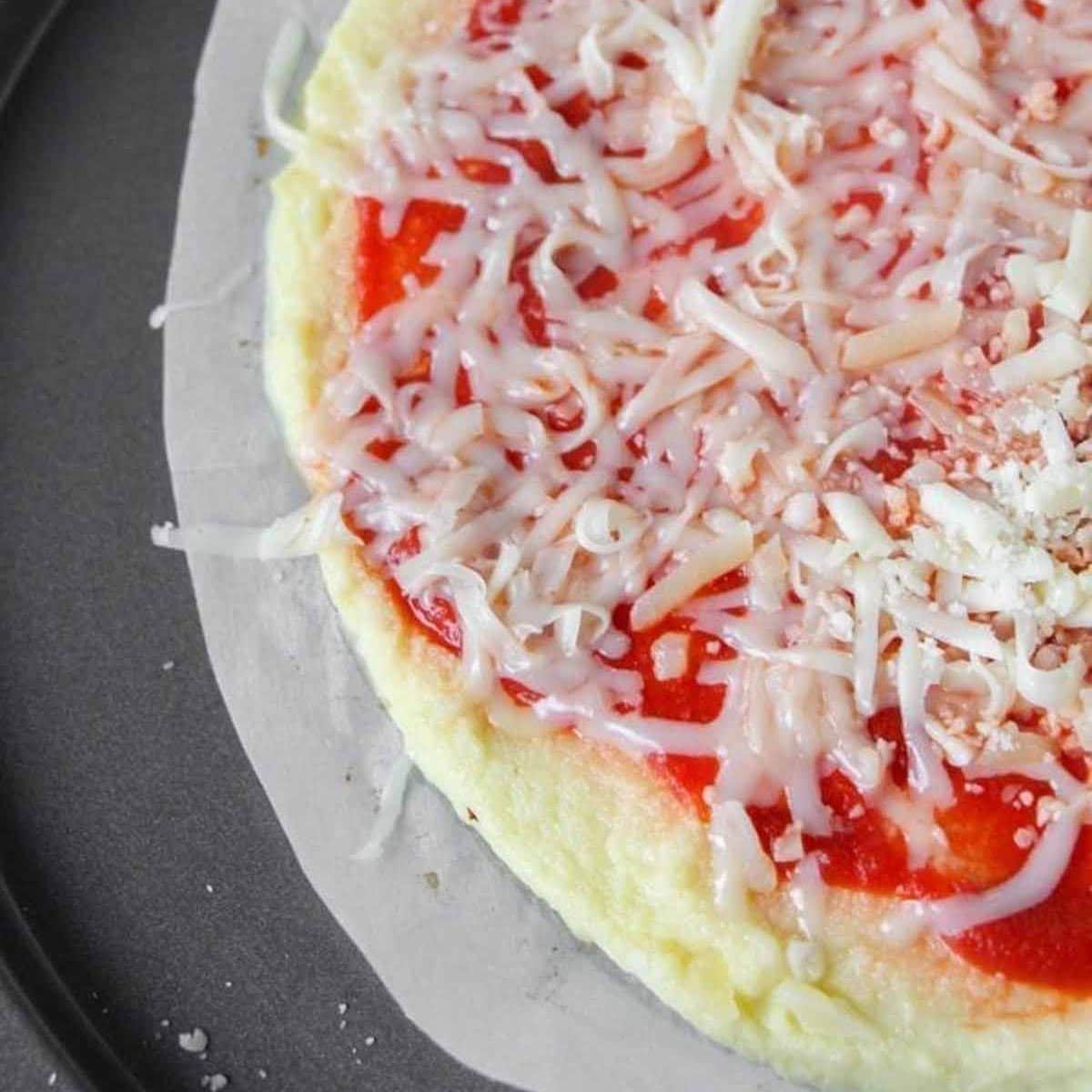 partially cooked cauliflower crust pizza with tomato based sauce and melting cheese on top of a parchment lined paper.