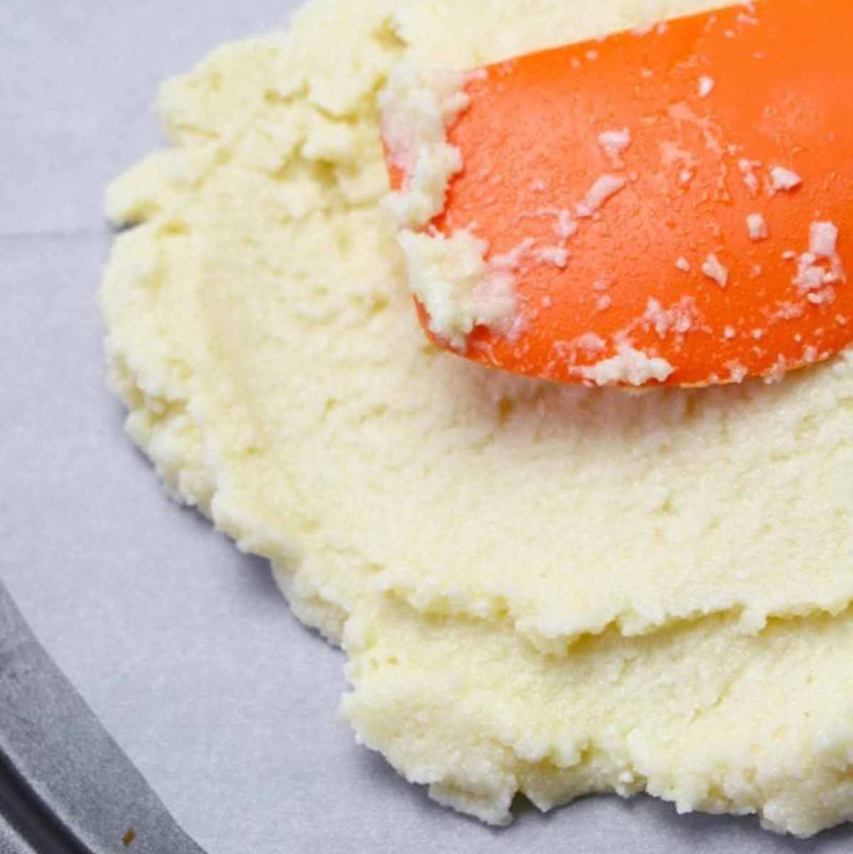 cauliflower, cheese and egg mixture being pressed on top a piece of parchment paper with an orange spatula.