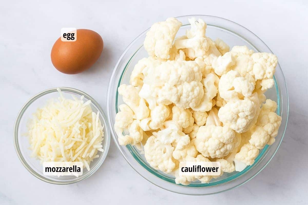 White counter with bowls of cauliflower and shredded mozzarella cheese and an egg.