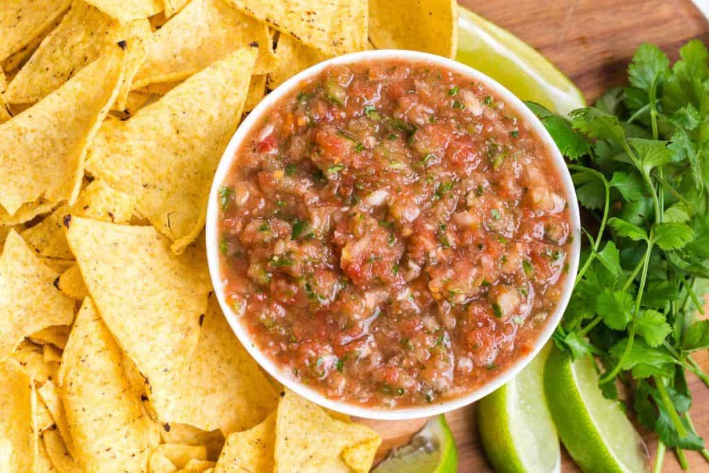 tortilla chips on left, fresh cilantro and lime wedges on the right with white bowl of salsa in the middle