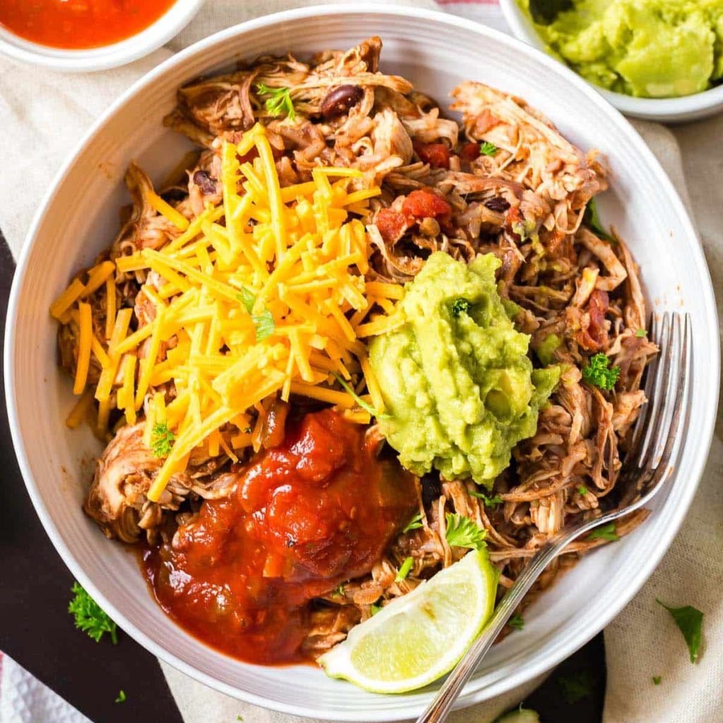 white bowl with chicken burrito bowls topped with shredded cheese, salsa, avocado and a lime wedge