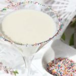 Martini glass rimmed with sprinkles and filled with white cocktail.