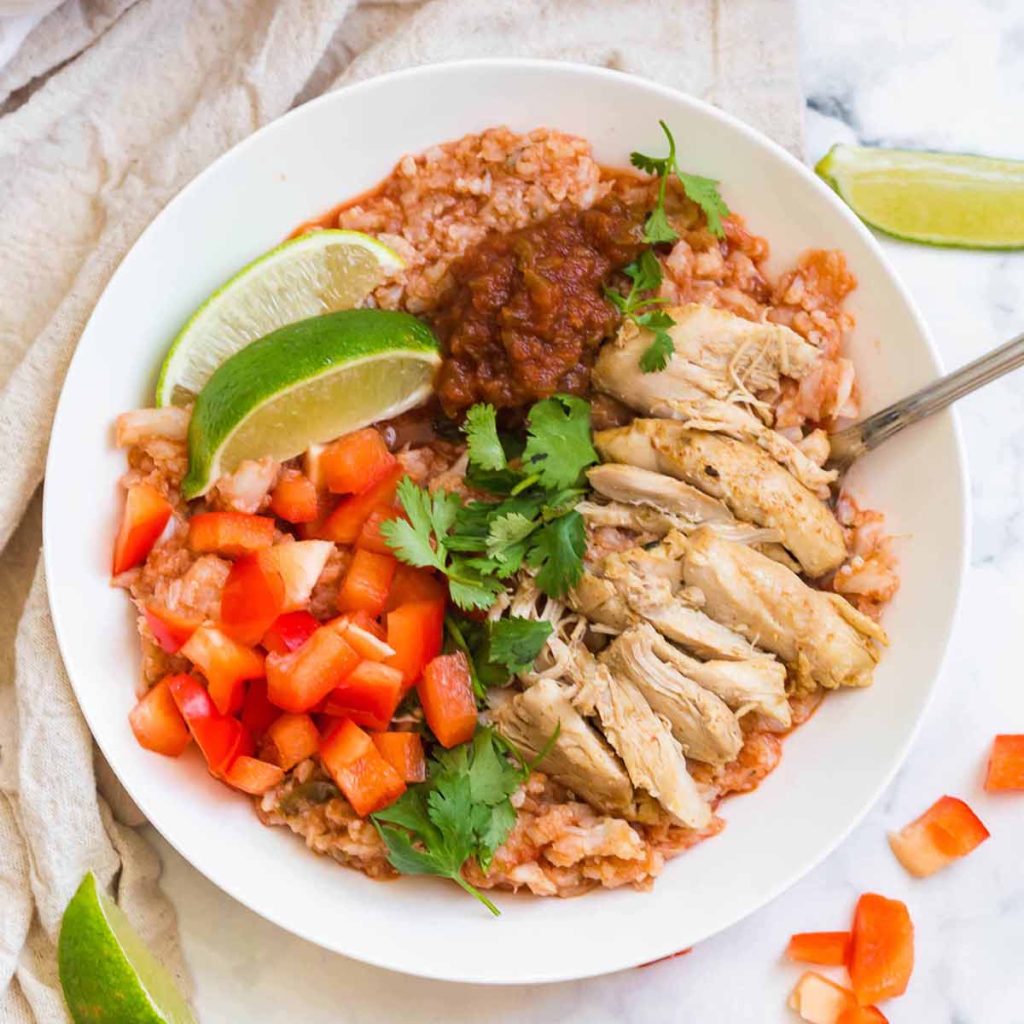 horizontal image of white bowl with cooked spanish cauliflower rice, chicken thighs, fresh tomatoes, and cilantro. There is a cream colored napkin in the background on the left side of the bowl.