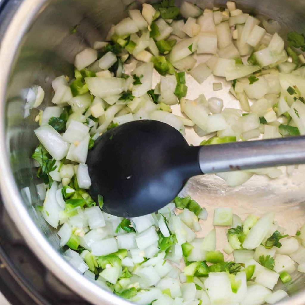 top down view of Instant Pot with onions, cilantro, and jalapeños cooking - there is a black spoon stirring the ingredients.