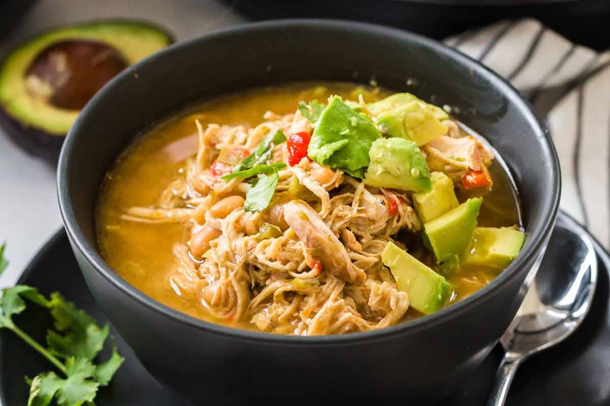A black bowl sitting on a white with black striped napkin. Inside the bowl is shredded chicken white chili that is topped with avocado pieces and cilantro.