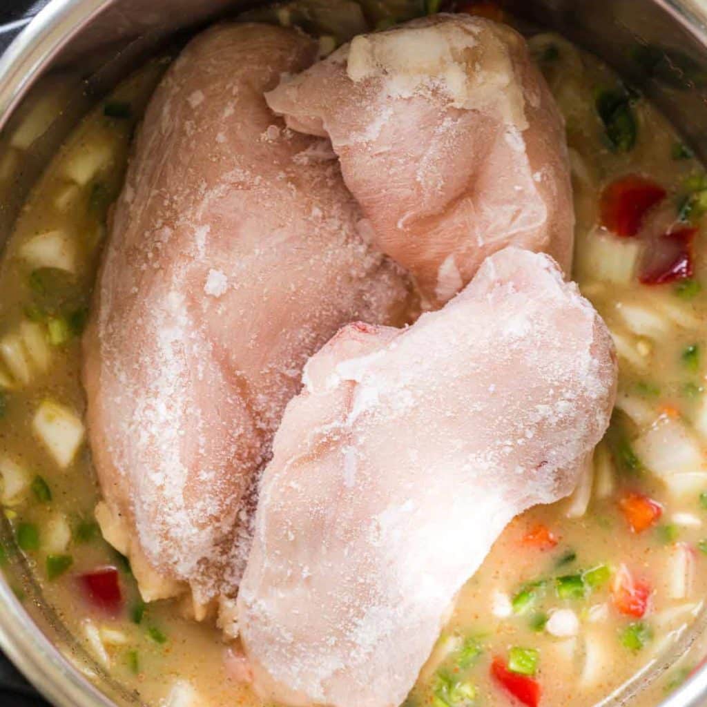 An instant pot with broth, vegetables, dry beans and three frozen chicken breasts sitting on top.