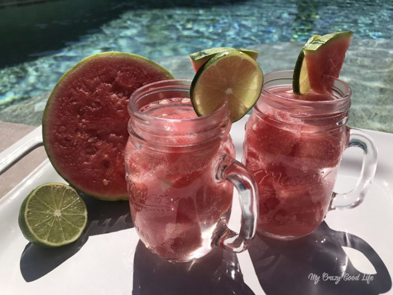 Two clear mason jars with handles and watermelon margaritas inside. There is frozen watermelon in the glass and slices of watermelon and lime on the rim. 