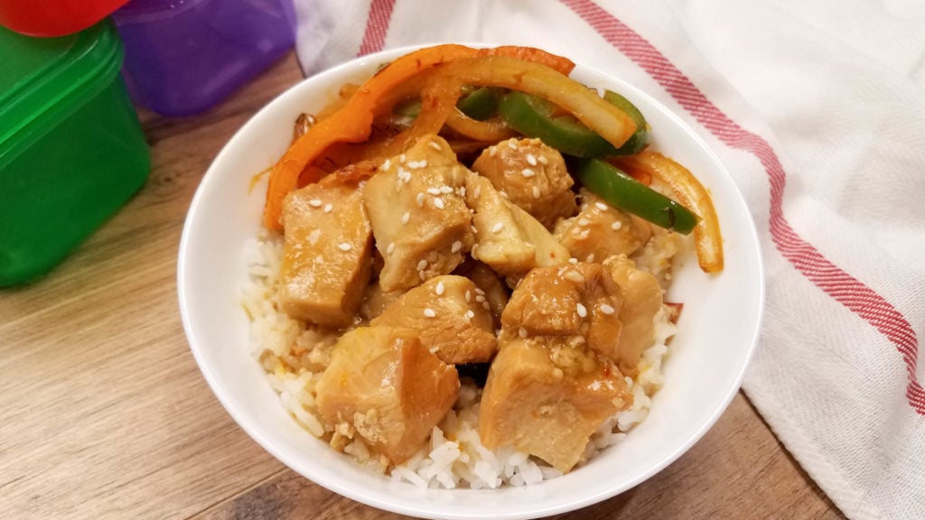 Orange chicken in a bowl on top of white rice with sautÃ©ed vegetables on the top side of the dish.