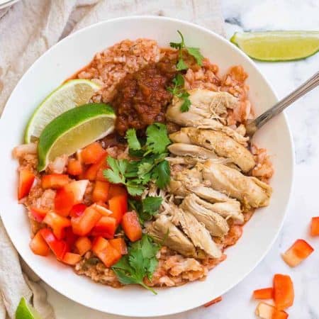 horizontal image of white bowl with cooked spanish cauliflower rice, chicken thighs, fresh tomatoes, and cilantro. There is a cream colored napkin in the background on the left side of the bowl.