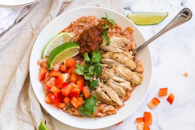 horizontal image of white bowl with cooked spanish cauliflower rice, chicken thighs, fresh tomatoes, and cilantro. There is a cream colored napkin in the background on the left side of the bowl.
