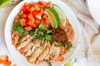 white bowl of cauliflower spanish rice and chicken thighs garnished with tomatoes, cilantro, salsa and two limes.