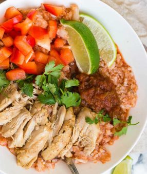 white bowl filled with cauliflower spanish rice and sliced chicken thighs garnished with salsa, cilantro, and limes.