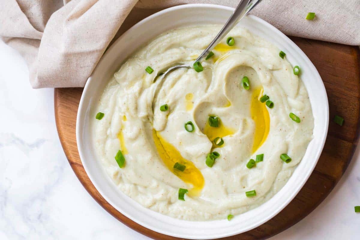 White bowl of mashed cauliflower with sliced green onions and ghee on top. 