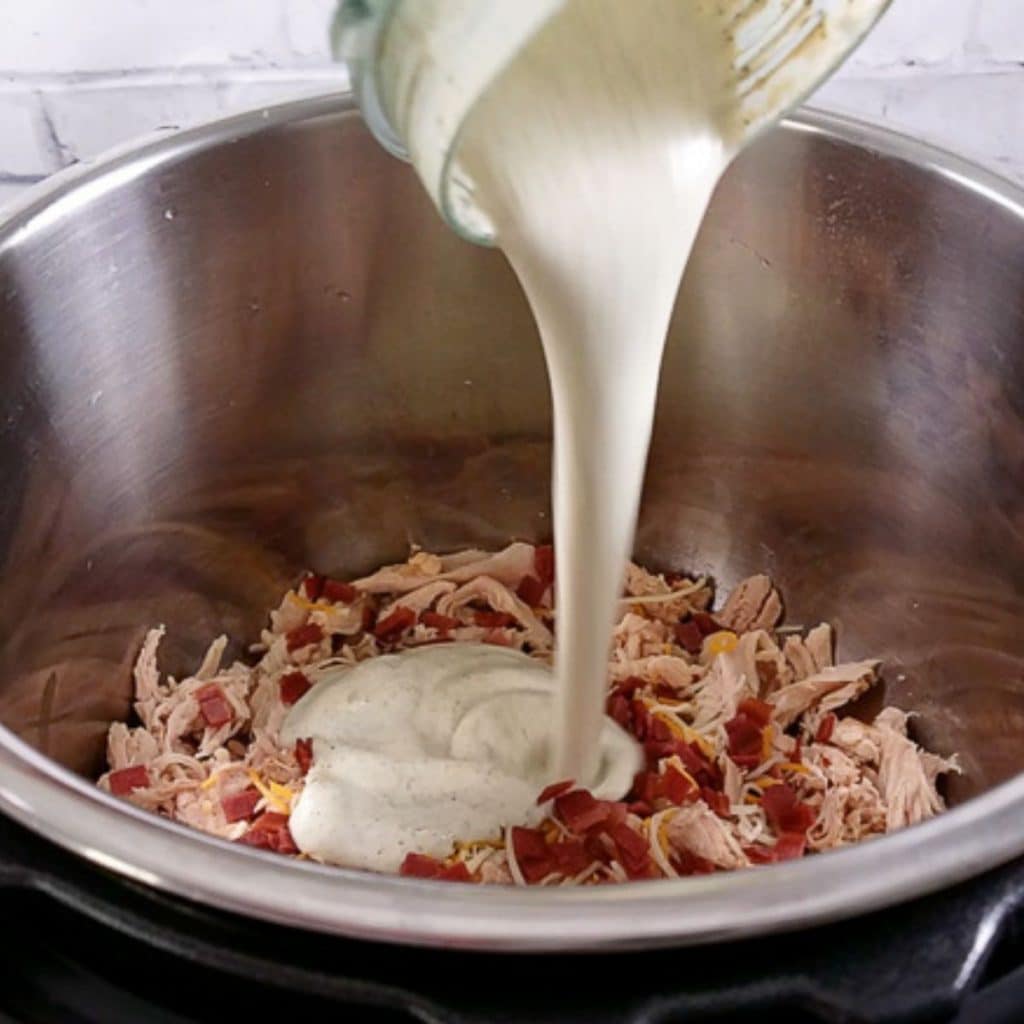 A jar of blended cottage cheese, greek yogurt and spices being poured into an instant pot full of shredded chicken and turkey bacon.