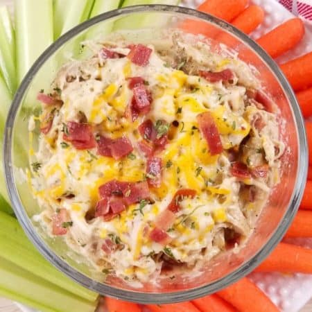 A glass bowl with shredded crack chicken with cheese and bacon, and surrounded by carrots and celery.