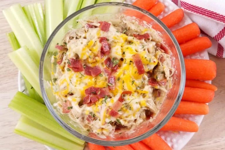 A glass bowl with shredded crack chicken with cheese and bacon, and surrounded by carrots and celery.