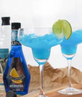 These frozen blue margaritas are delicious and simple! Give them a try today. This is a photo of the blue margarita mix frozen and placed in a glass with the ingredients in the background.