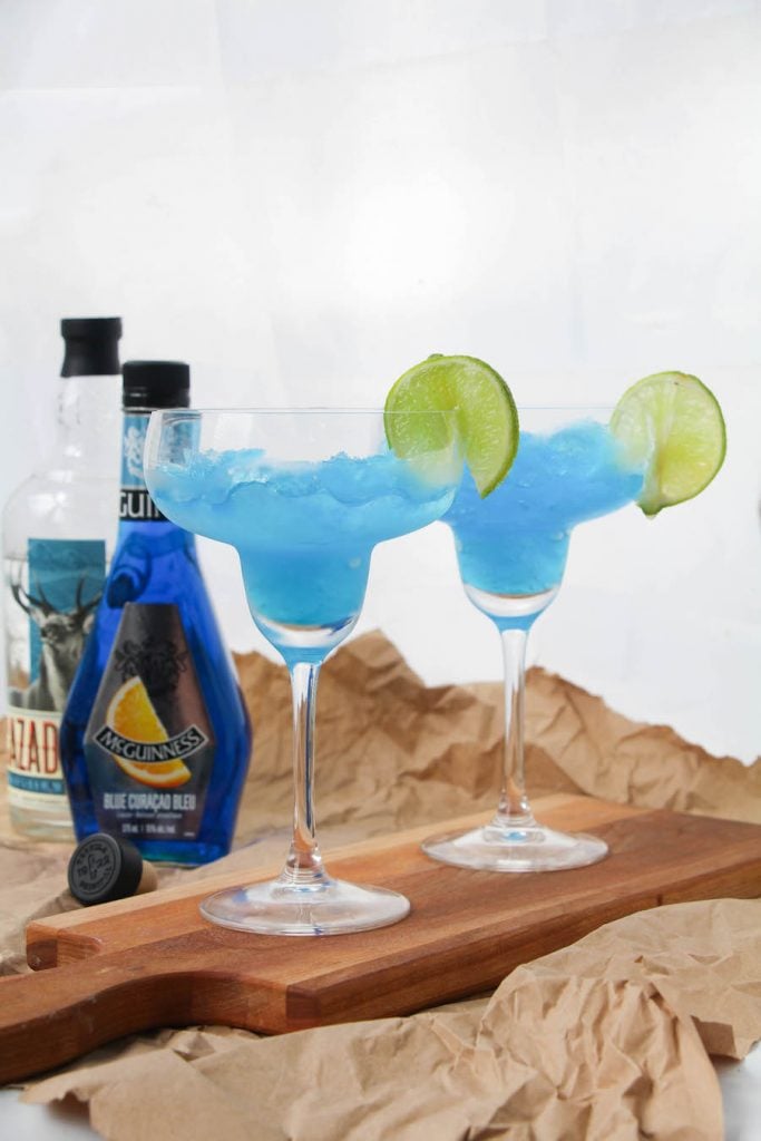 Two frozen blue margaritas in classic margarita glasses with a lime slice. 